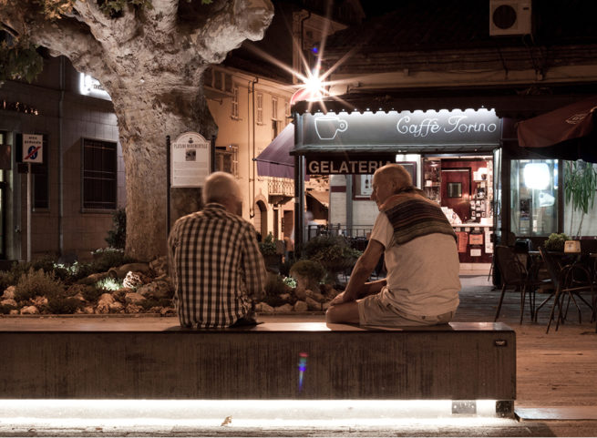 Due uomini seduti su una panchina davanti al Caffè Torino illuminato di sera.