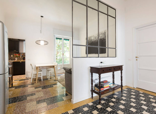 Interno luminoso con pavimento in graniglia, tavolo da pranzo e divisorio in vetro industriale.